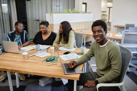 Portrait of smiling handsome young Black man attending business meeting and working on laptop