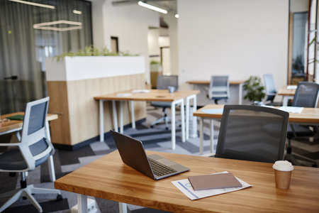 Desk with laptop, cup of coffee and report in empty office in modern building