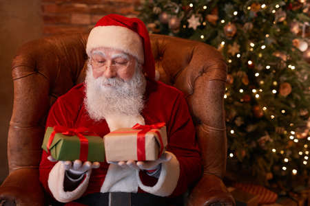 Thoughtful Santa Claus in costume sitting in comfortable armchair against Christmas tree and choosing giftの写真素材