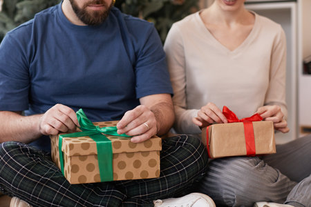 Close-up of young couple sitting with crossed legs and unwrapping Christmas gifts at same timeの写真素材