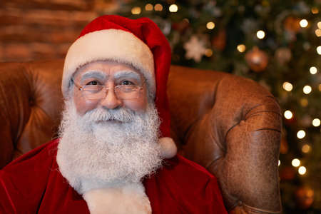Portrait of content senior Santa Claus in red cap and eyeglasses sitting in armchair against Christmas treeの写真素材