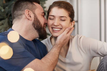 Happy young bearded man kissing beloved woman with closed eyes on New Year, lens flare effect
