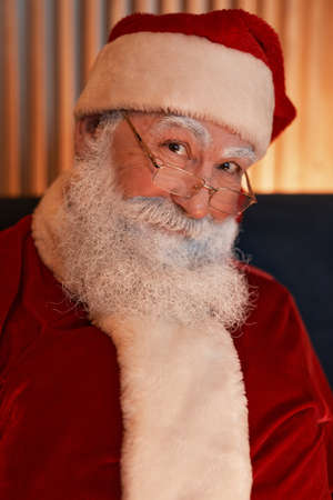 Portrait of smiling kind senior white-bearded Santa Claus in cap and eyeglasses wearing red costume