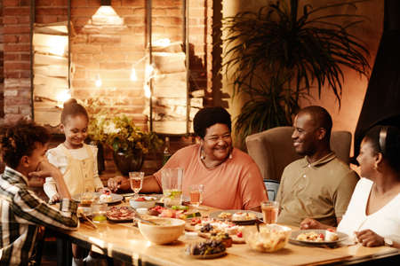 Warm toned portrait of big African-American family sitting at table outdoors and enjoying dinner together at eveningの写真素材
