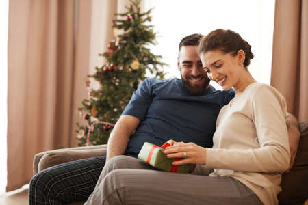 Smiling young woman sitting on sofa in living room with Christmas tree and unpacking present from boyfriendの写真素材