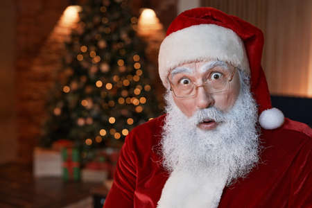 Portrait Of surprised Santa Claus in eyeglasses keeping mouth open in room with Christmas treeの写真素材
