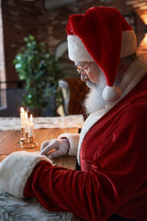 Serious Santa Claus in red cap and glasses sitting at wooden table with burning candles and reading letterの写真素材