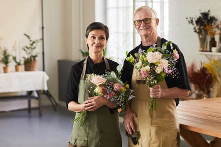 Waist up portrait of mature couple holding flowers in flower shop and looking at cameraの写真素材