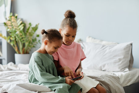 Portrait of two cute African-American girls using smartphone together while sitting on bed in cozy room, copy spaceの写真素材