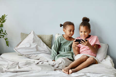 Front view portrait of two cute African-American girls using smartphone together while sitting on bed in cozy room, copy spaceの写真素材