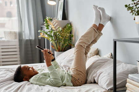 Full length portrait of teenage African-American boy using smartphone on bed while lying feet up, copy spaceの写真素材