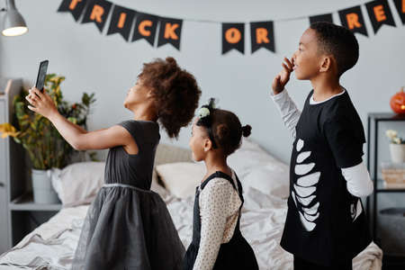 Side view portrait of cute African-American children wearing Halloween costumes at home and waving at camera while video chatting with familyの写真素材