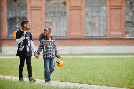Full length portrait of two African-American boys holding hands and wearing Halloween costumes while trick or treating together, copy space