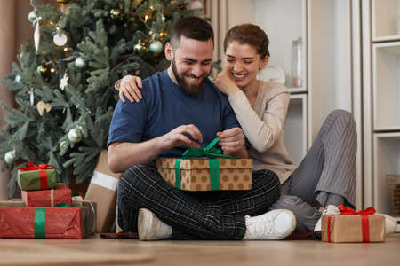 Smiling young bearded man embraced by girlfriend sitting with crossed legs against Christmas tree and unwrapping presentの写真素材