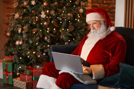 Concentrated aged Santa Claus sitting on sofa near Christmas tree and working with laptop