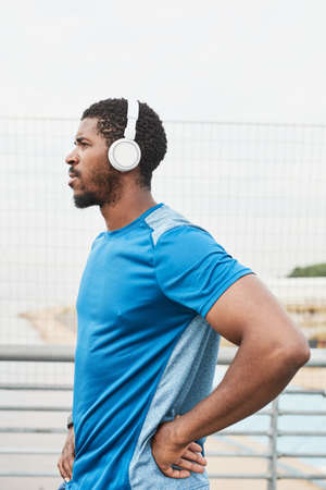 Side view of African young athlete in wireless headphones listening to music during training...