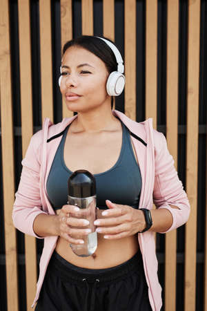 African beautiful woman in wireless headphones holding bottle of water during training outdoorsの写真素材