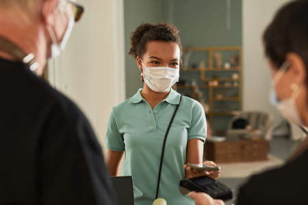 Portrait of young female customer paying via smartphone in flower shop and wearing mask, copy space