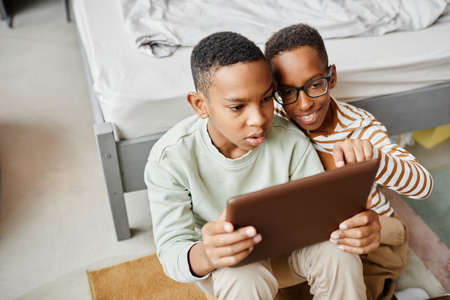 High angle view at two teenage African-American boys using digital tablet together while siting on floor in cozy room, copy spaceの写真素材