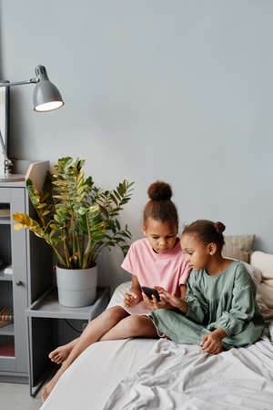 Vertical full length portrait of two African-American girls using smartphone together on bed in cozy room, copy spaceの写真素材