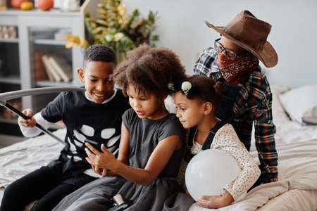 Group of African-American kids wearing Halloween costumes at home and using smartphoneの写真素材
