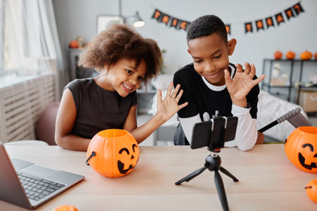 Two smiling African-American kids waving at smartphone while video chatting on live streaming on Halloweenの写真素材