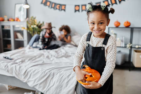 Waist up portrait of happy African-American girl holding carved pumpkin and smiling at camera while standing in room with Halloween decorations, copy spaceの写真素材
