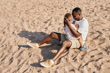 Full length portrait of young African-American father holding son while sitting on sand at beach together, copy spaceの写真素材