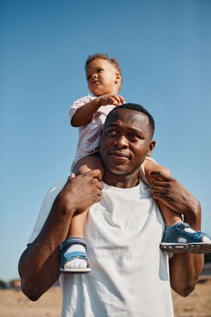 Vertical portrait of happy African-American father carrying son on shoulders against blue sky while enjoying walk on beachの写真素材