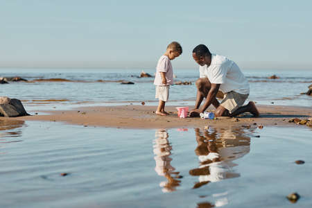 Minimal side view portrait of young African-American father and son playing with sand on beach together, copy spaceの写真素材