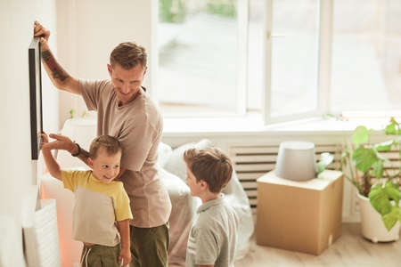 Side view portrait of happy father with two sons hanging pictures on wall while moving in to new home, copy spaceの写真素材