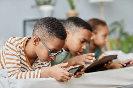 Side view portrait of three kids in row using gadgets while lying on bed at home, copy spaceの写真素材