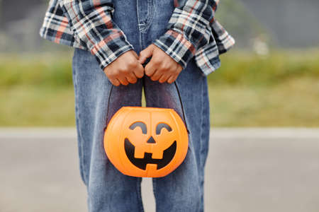 Close up of unrecognizable boy holding pumpkin shaped Halloween pail outdoors, copy space