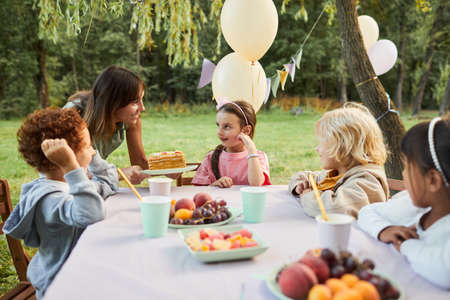 Portrait of young mother bringing Birthday cake to smiling girl during Birthday party outdoors, copy...