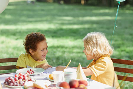 Two little kids at picnic table outdoors decorated with balloons for Birthday party in Summer, copy spaceの写真素材