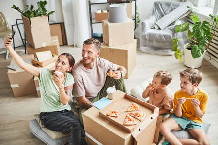Portrait of happy family eating pizza from cardboard box and taking selfie photo while celebrating moving in to new house, copy spaceの写真素材