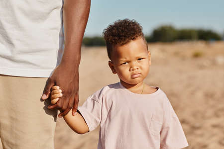 Portrait of cute African-American toddler holding hands with dad while enjoying walk on beach in sunlightの写真素材
