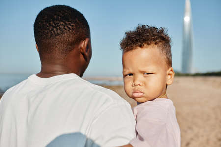 Portrait of cute African-American baby looking at camera while enjoying walk on beach in fathers arms, copy spaceの写真素材