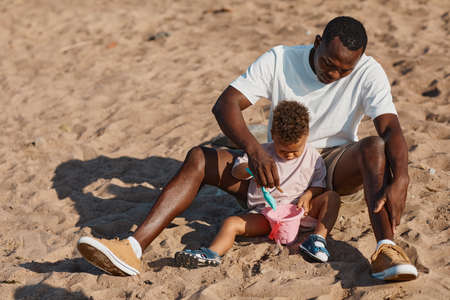 High angle portrait of young African-American father and son playing in sand together at beach, copy spaceの写真素材