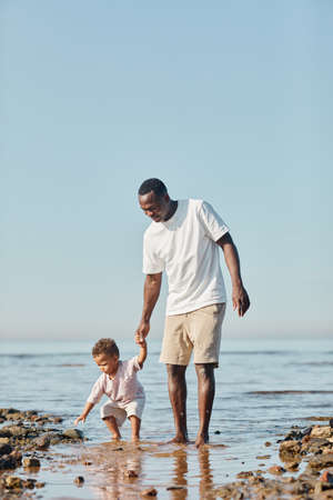Vertical full length portrait of happy young father and son enjoying walk on beach together and playing in waterの写真素材