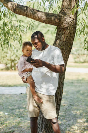 Vertical portrait of African-American man holding cute baby son in park while using smartphoneの写真素材