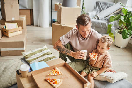Portrait of happy father and son eating pizza from cardboard box while celebrating moving in to new house, copy spaceの写真素材