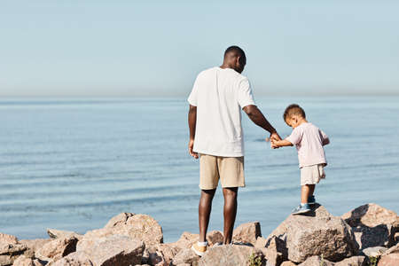 Minimal back view portrait young African-America father holding cute son while playing on beach together, copy spaceの写真素材