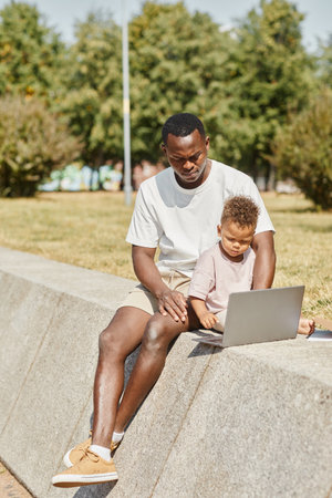 Vertical full length portrait of young African-American man using laptop outdoors with cute baby sonの写真素材