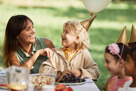 Portrait of happy young woman with son sitting at picnic table with group of kids during outdoor Birthday party in Summerの写真素材
