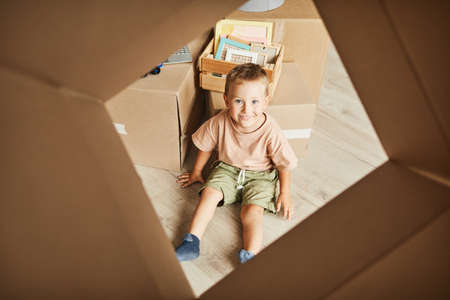 From the box shot of cute boy sitting on floor and playing with cardboard boxes while family moving to new house, copy spaceの写真素材