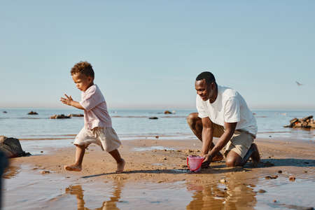 Side view portrait of happy African-American father playing with baby boy running on beach, copy spaceの写真素材