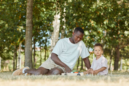 Portrait of young African-American father playing with cute son in park while sitting on grass, copy spaceの写真素材