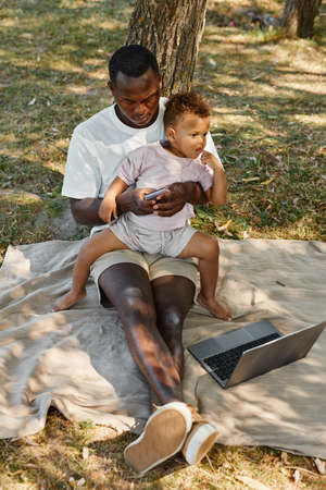 Vertical full length portrait of African-American man using laptop outdoors in park with cute baby son, copy spaceの写真素材