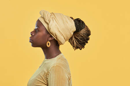 Minimal side view portrait of young African-American woman wearing head wrap and ethnic jewelry while standing against yellow background in studio, copy spaceの写真素材